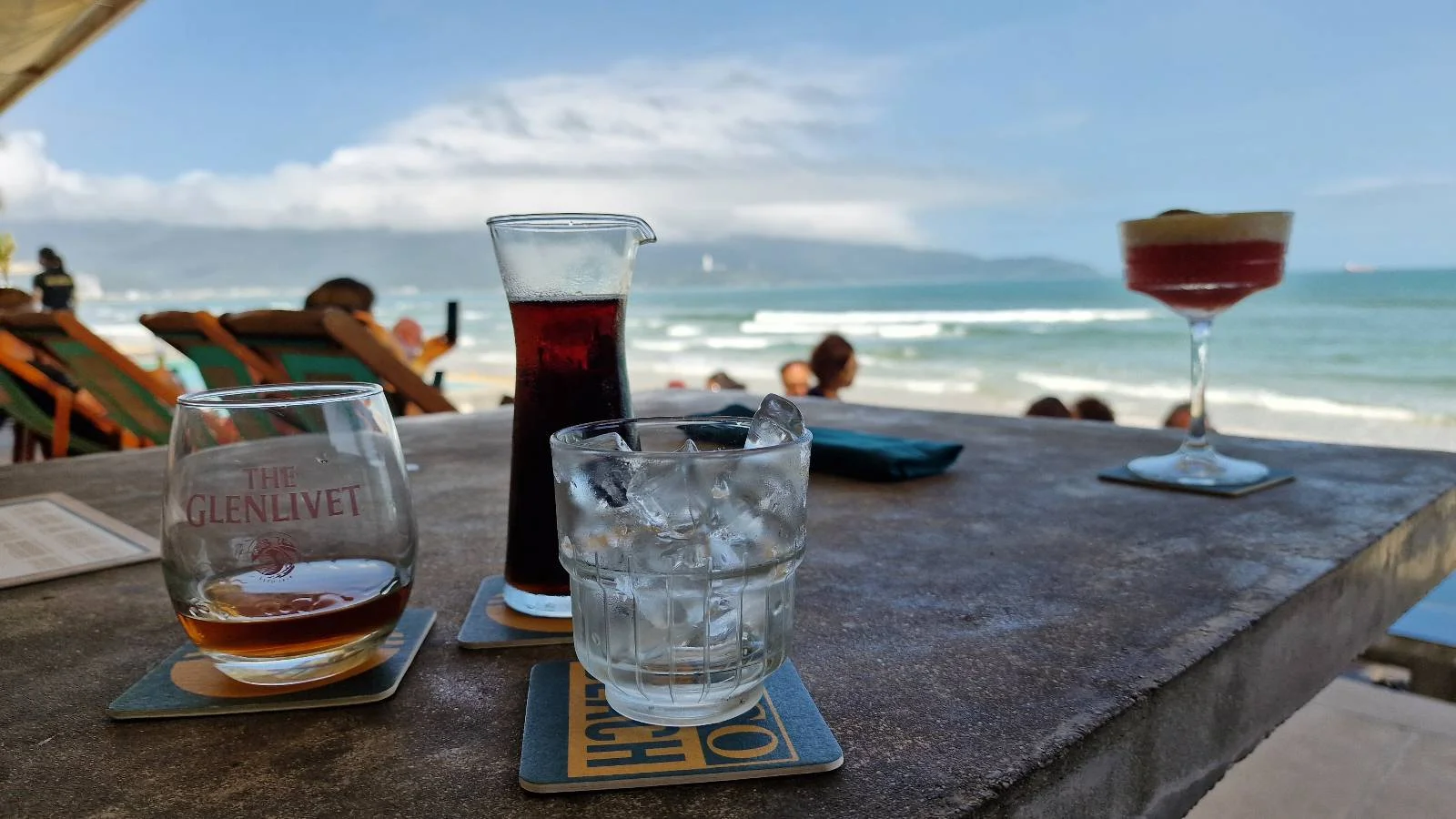 Three drinks—one with ice, one dark, and one red cocktail—sit on a table overlooking a beach with people relaxing on lounge chairs and waves in the background under a partly cloudy sky.