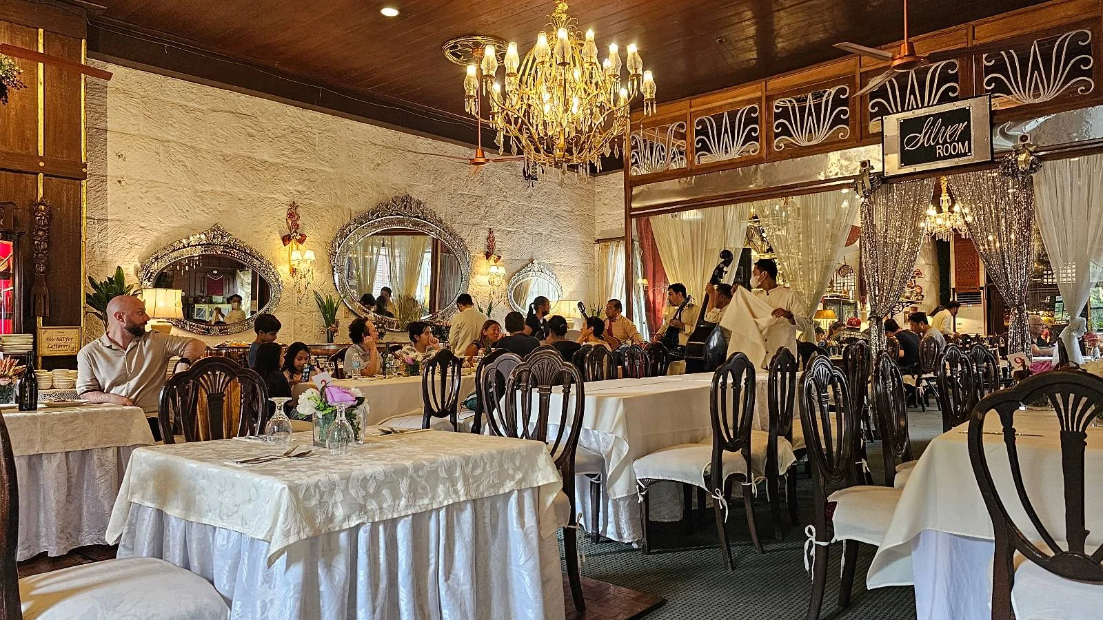 A restaurant interior with people dining at tables covered with white tablecloths. Decorative mirrors and wooden furniture add to the elegant atmosphere. Chandeliers hang from the ceiling.