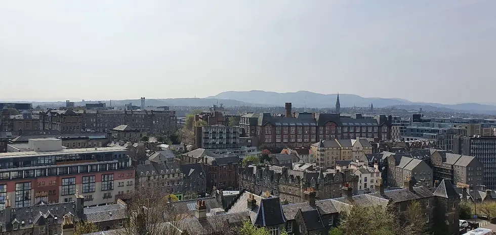 Cityscape with historic stone buildings and modern structures under a clear sky; distant hills on the horizon create a serene mood.