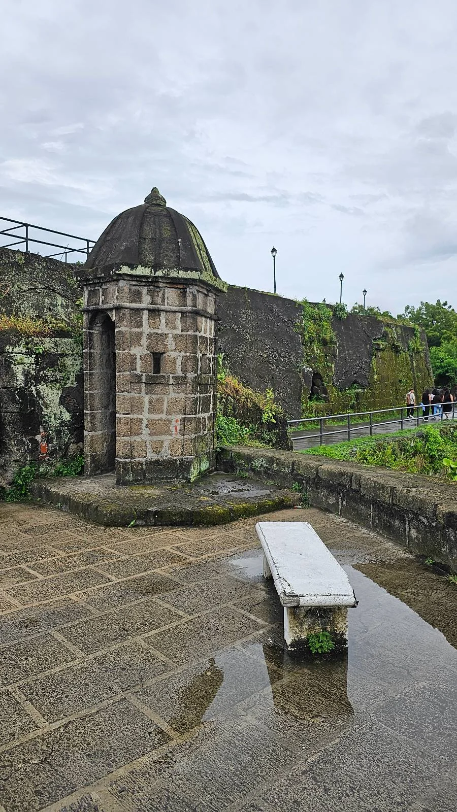 A small stone tower with a conical roof stands next to an ancient stone wall, covered in patches of greenery. In front, a narrow stone bench rests on a wet patio. The sky is overcast, creating a serene and historical atmosphere.