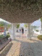 Woman in shorts walks along a decorative path under intricate archways, surrounded by sculptures and greenery. Bright, sunny day.