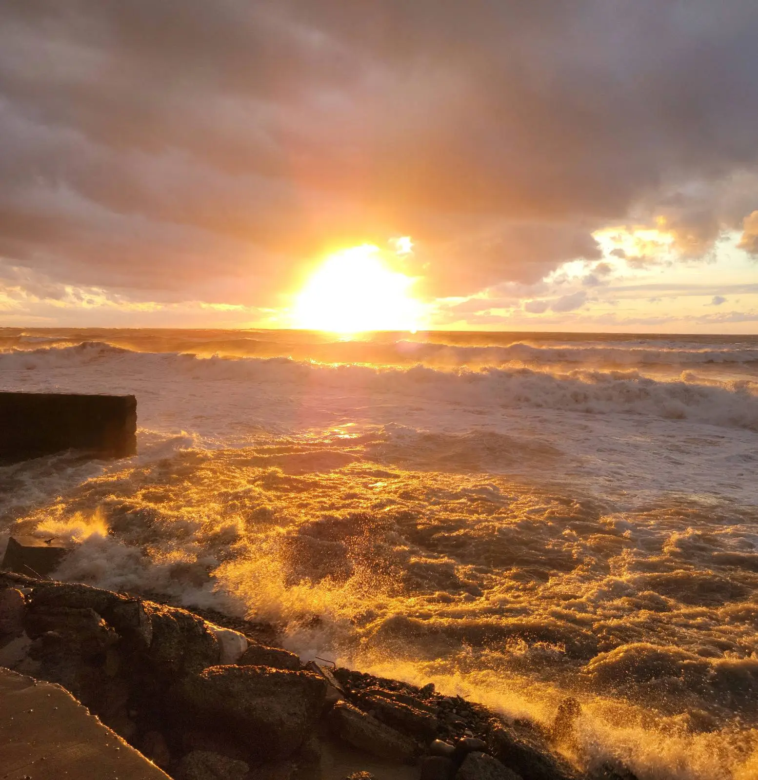A dramatic sunset over a turbulent ocean, with waves crashing onto rocks in the foreground and golden sunlight reflecting off the water under a cloudy sky.