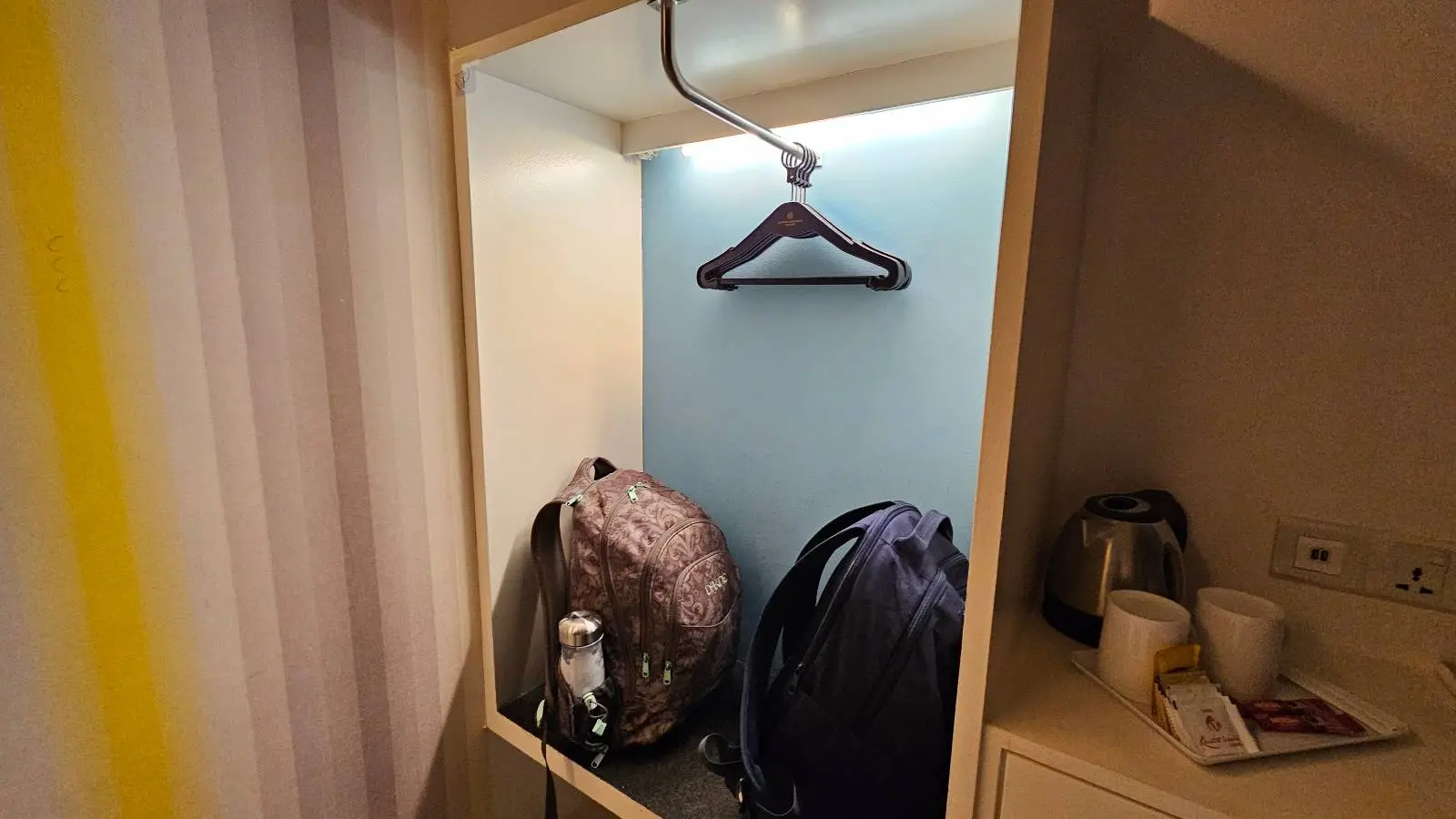 Open wardrobe with a single dark clothes hanger, a patterned backpack, a black bag, and a water bottle on the shelf. To the right, there is an electric kettle and two cups on a small counter.