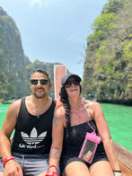A man and a woman sit on a boat in a tropical lagoon, embodying the spirit of slow travel. With turquoise waters and lush green cliffs around them, they bask in serenity—him in a tank top, her in a black sun hat and dress—both smiling behind their sunglasses.