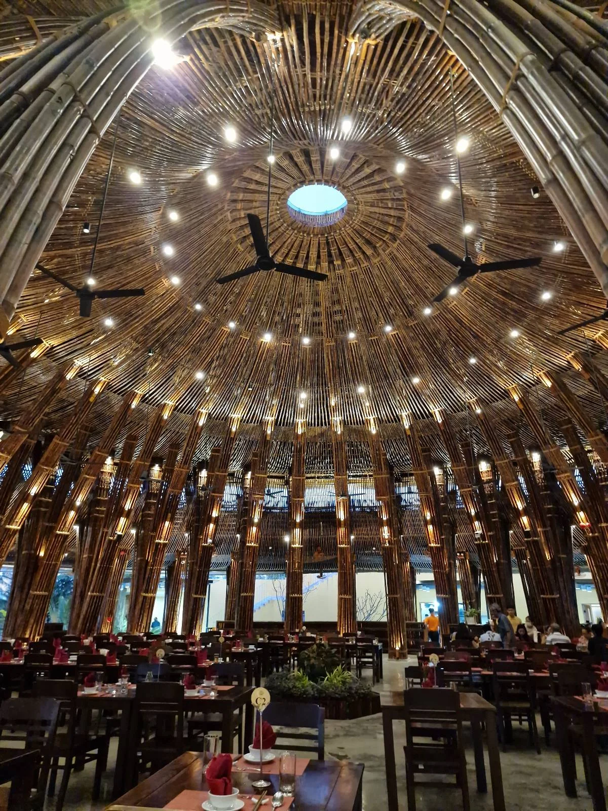 Interior of a restaurant featuring a high ceiling made of bamboo sticks arranged radiating from the center, with hanging lights and ceiling fans, furnished with dark tables and red chairs.
