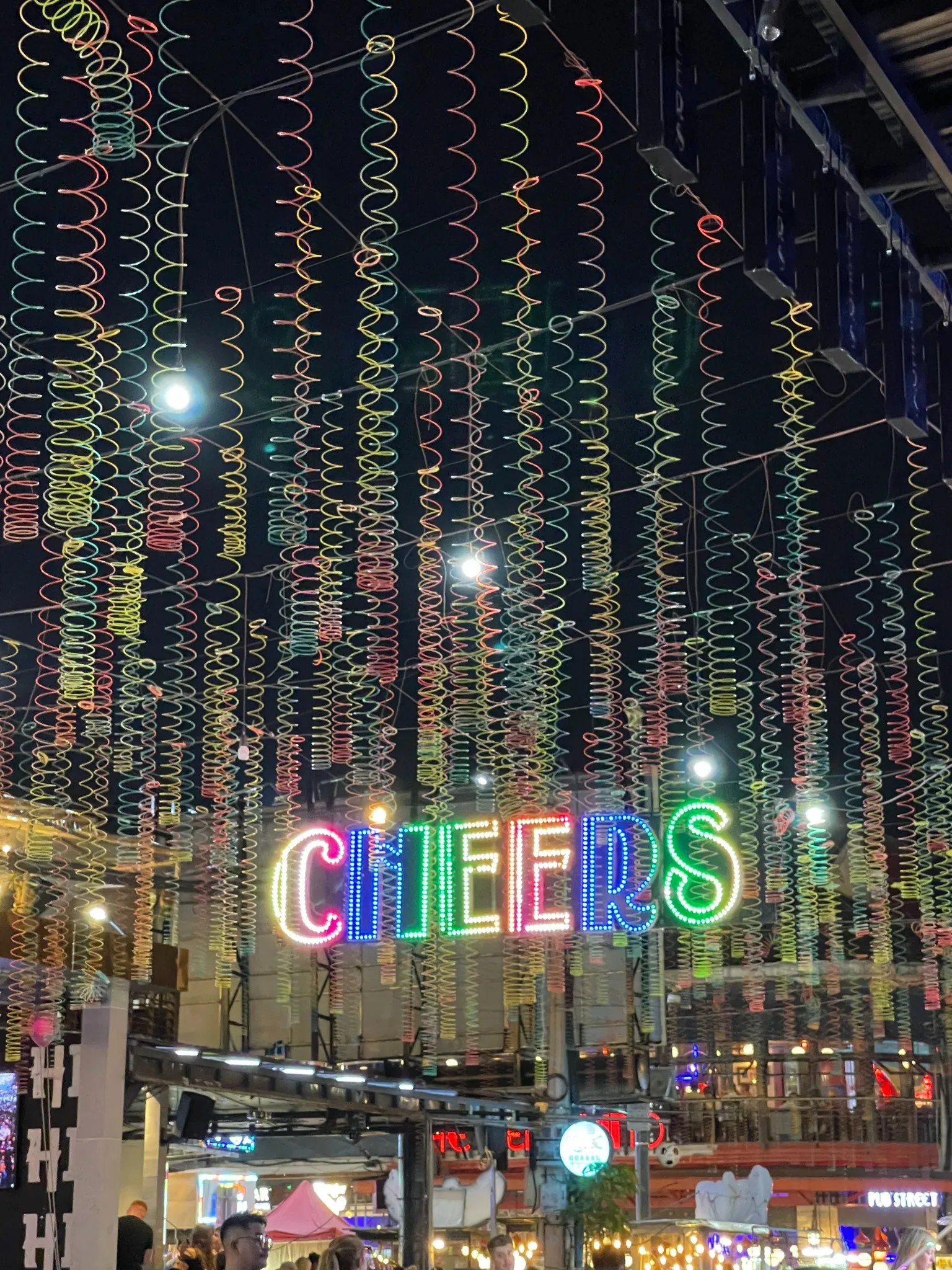 Colorful neon sign spelling "CHEERS" hangs above a lively indoor space, with spiral decorations and string lights overhead, creating a festive atmosphere. People can be seen gathered below.
