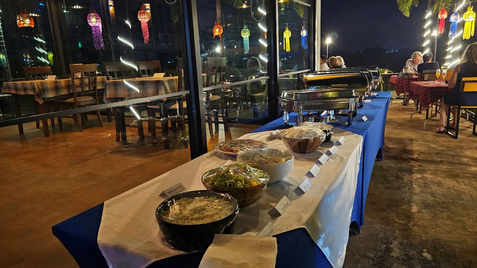 Buffet table with salad bowls and food trays on a blue and white tablecloth at an outdoor restaurant with string lights and lanterns.