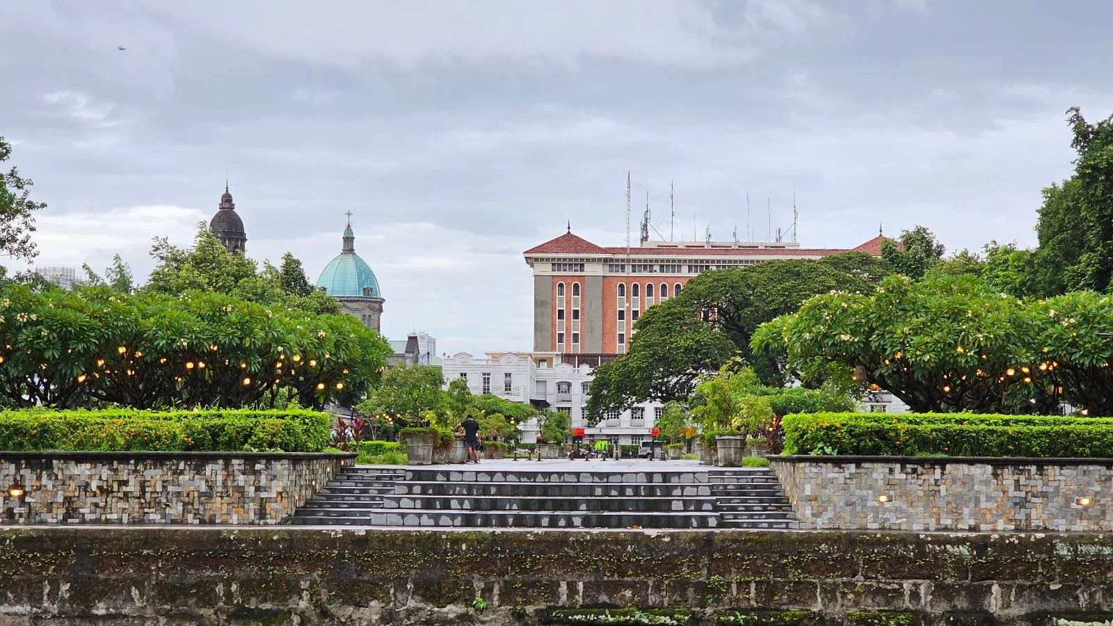 A beautifully manicured park with a cascading fountain in the foreground. Green trees and shrubs line the path. In the background, a historic building with a domed roof and another with red brick architecture are visible under an overcast sky.