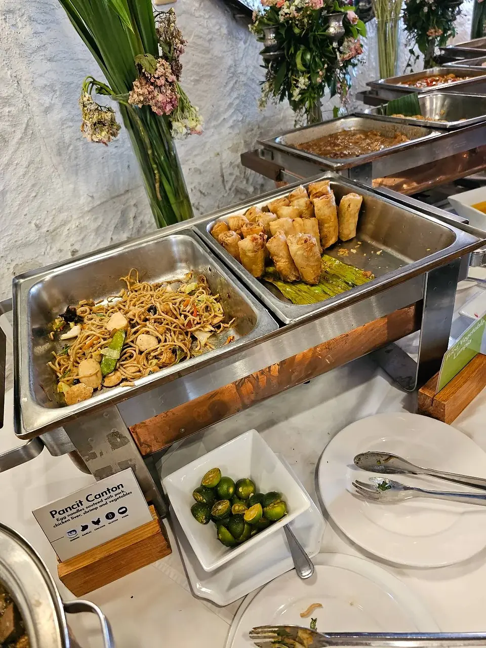 Buffet table with trays of noodle stir-fry and fried spring rolls, garnished with lemongrass. A small bowl of limes and serving utensils are placed nearby.