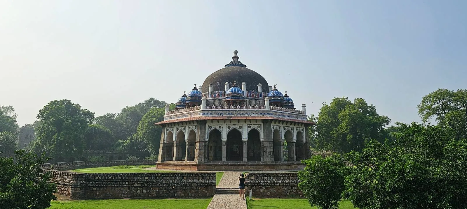 A panorama of Isa Khan's Tomb, an ornate, octagonal mausoleum with arches and a large dome, surrounded by green lawns and trees. The sky is clear and blue, enhancing the historic architecture.