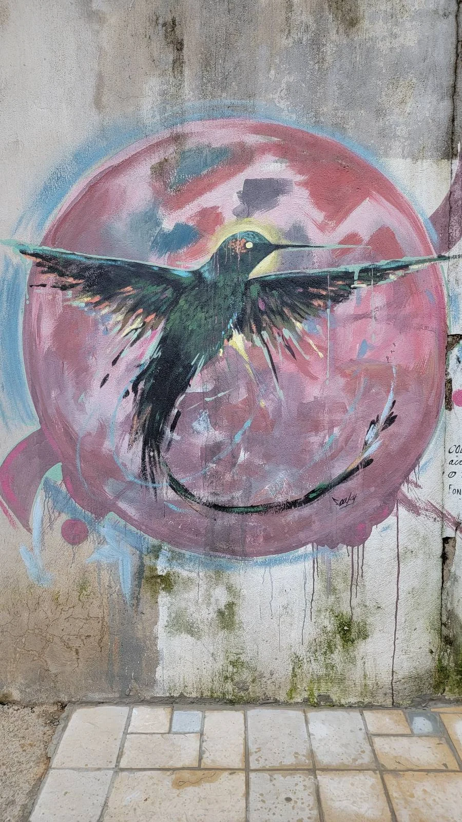 A mural on a weathered wall depicting a large bird with outstretched wings set against a circular, colorful, abstract background. The scene shows artistic splashes of pink, blue, and green, adding a dynamic feel to the composition.