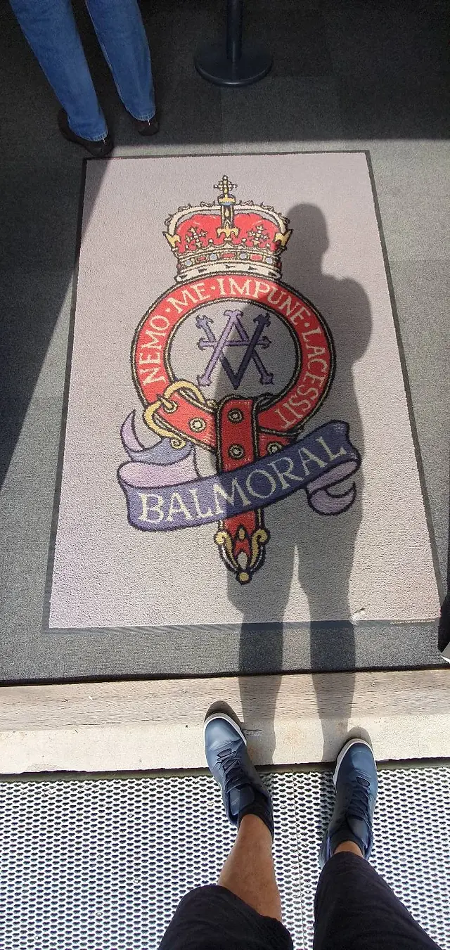 A person stands at the entrance of a building, casting a shadow over a doormat. The doormat features a crest with a crown, a lion, and the word "BALMORAL" on it. The person is wearing black shoes and shorts.