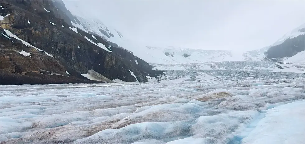 Snow-covered glacier with rocky mountains in the background under an overcast sky.