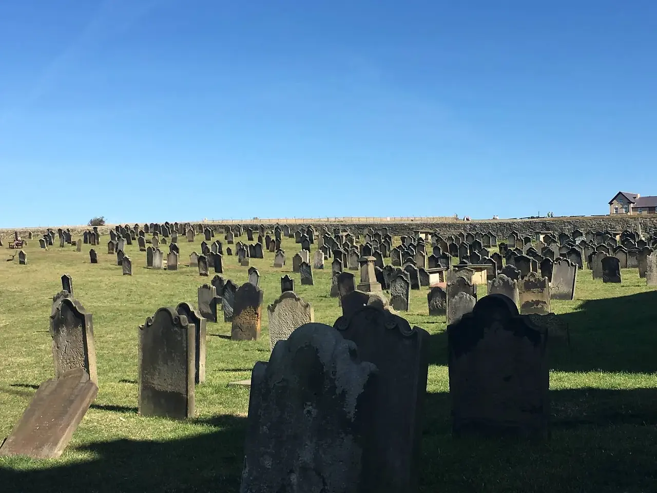 A grassy hillside cemetery with numerous old, weathered tombstones under a clear blue sky. The gravestones are arranged in rows, casting long shadows on the grass. A small building is visible in the distance.