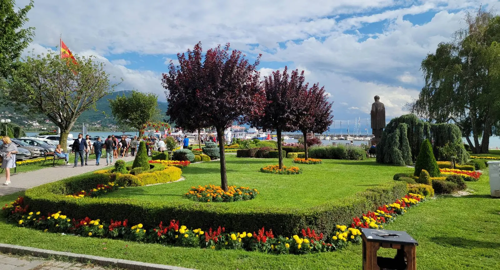 A scenic park with neatly trimmed grass, flower beds, and several trees. People walk along a path on the left. In the background, there is a lake and distant mountains under a partly cloudy sky.