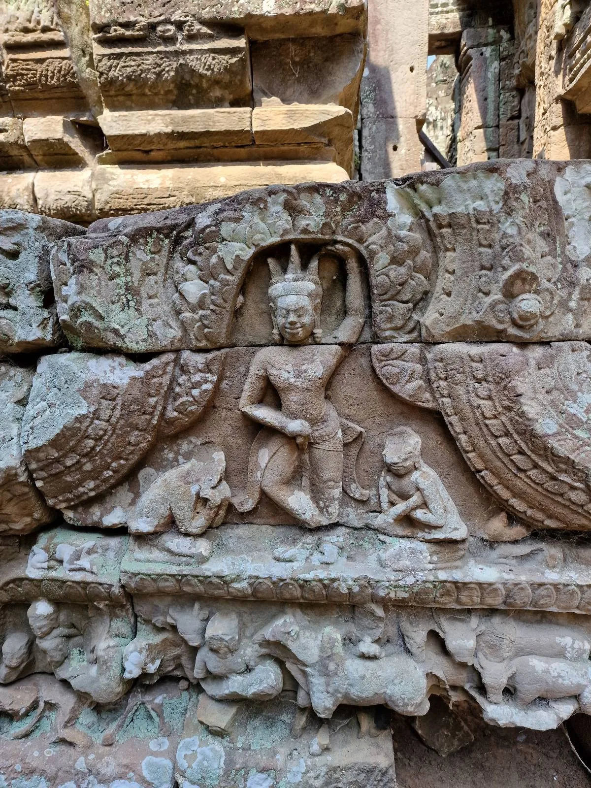 Ancient stone carving depicting a crowned figure seated in the center, surrounded by animals and other human figures, with intricate detailing and weathered textures on the temple wall.