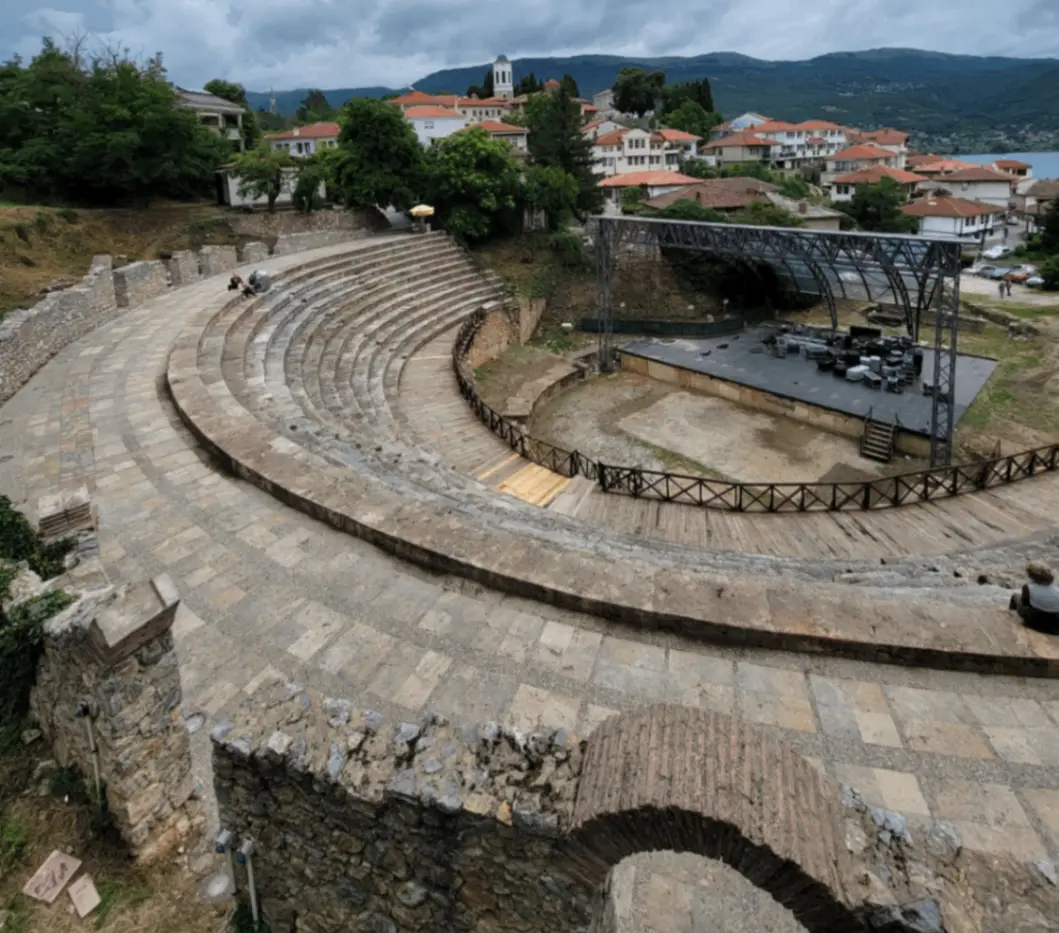 An ancient stone amphitheater with curved seating, set outdoors near a small town. There are modern buildings and lush trees in the background under a cloudy sky.