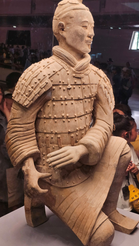 A clay statue of a kneeling ancient Chinese warrior in detailed armor, part of the Terracotta Army, is displayed indoors with people and dim lighting in the background.