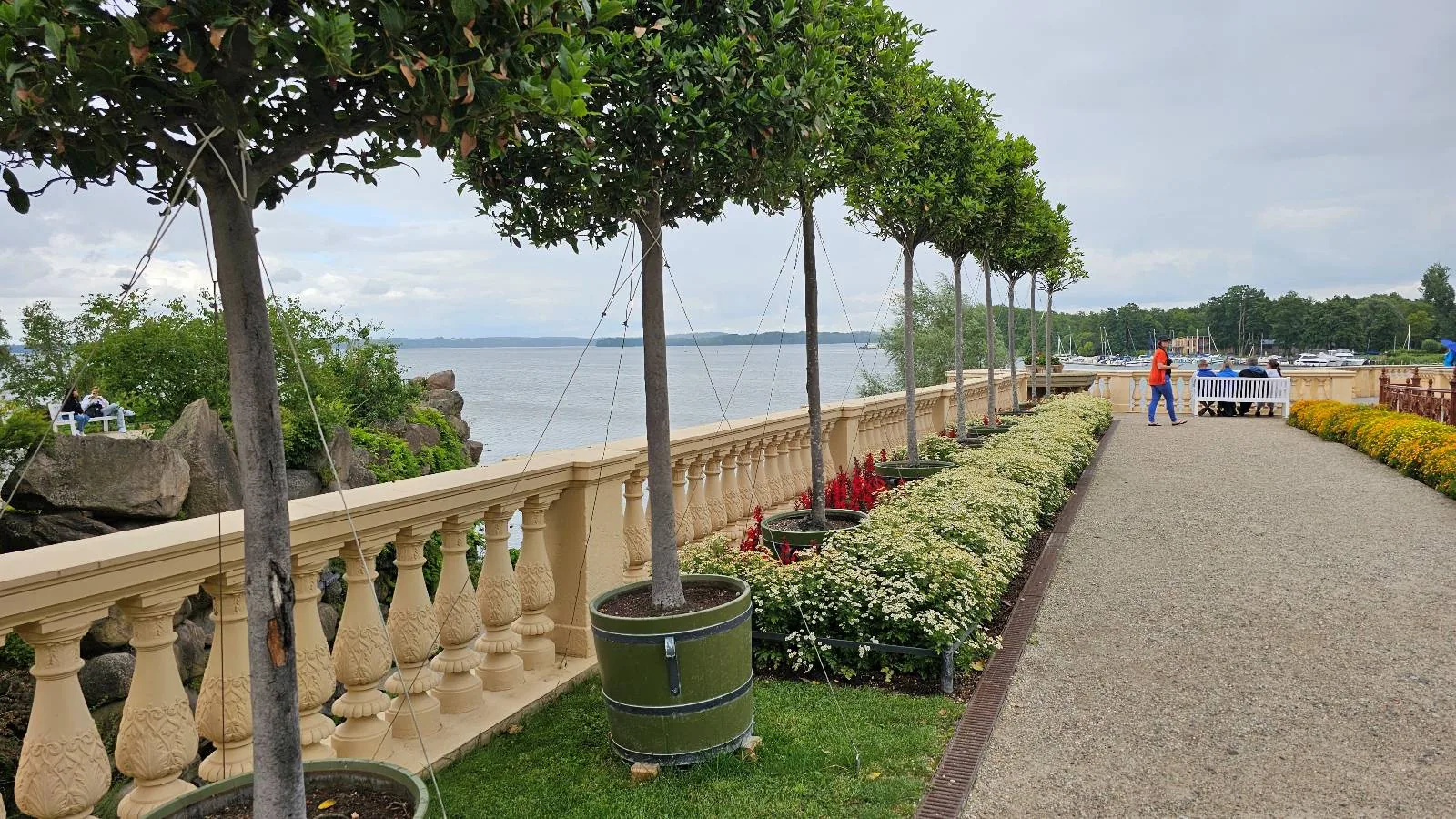 A paved walkway lined with trimmed trees and flowering bushes runs alongside a stone railing overlooking a body of water. Two people are visible in the distance near parked cars.