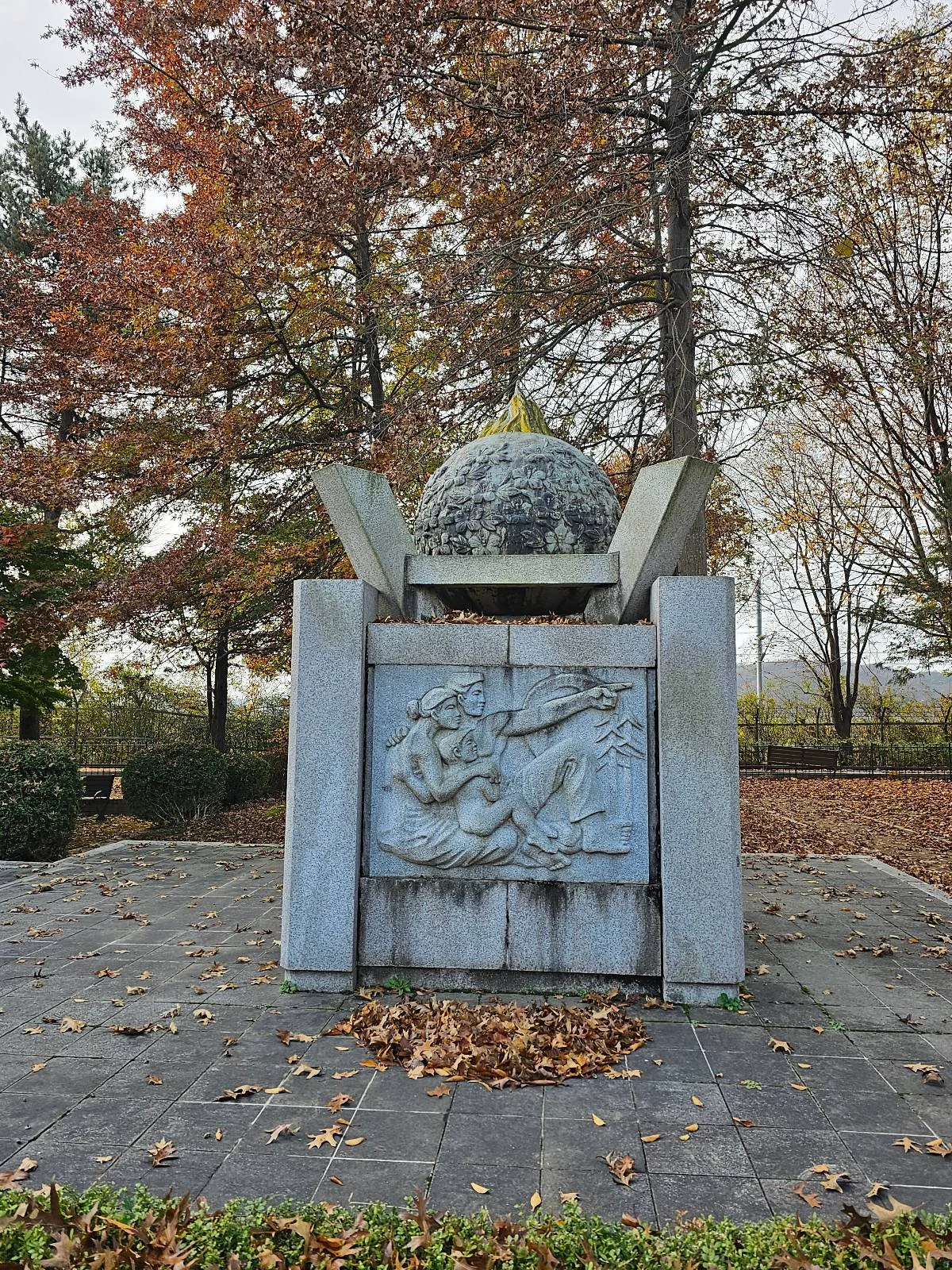 A monument features a globe atop a square base with engraved figures. It stands on a stone platform surrounded by fallen autumn leaves and trees with colorful foliage. The sky is overcast, adding a somber atmosphere to the scene.