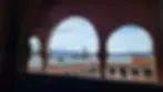 View through arched openings showcasing a distant landscape and a building with a terracotta-tiled roof.