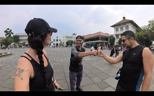 Three digital nomads in casual attire are fist bumping in an open plaza, surrounded by historic buildings. The person on the left showcases a tattoo and braided hair. It's a sunny day, embodying the slow travel lifestyle and bringing lives closer together amidst rich history.