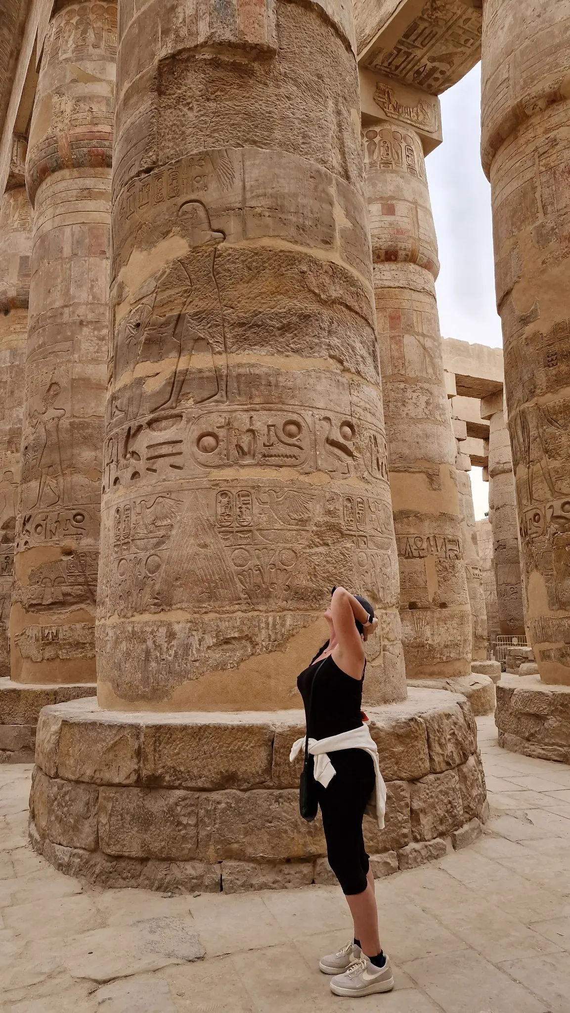 A person in black workout clothes and a white shirt tied around their waist stands with one arm raised, gazing up at a massive ancient stone column, with more columns visible in the background. The setting appears to be historical and outdoors.