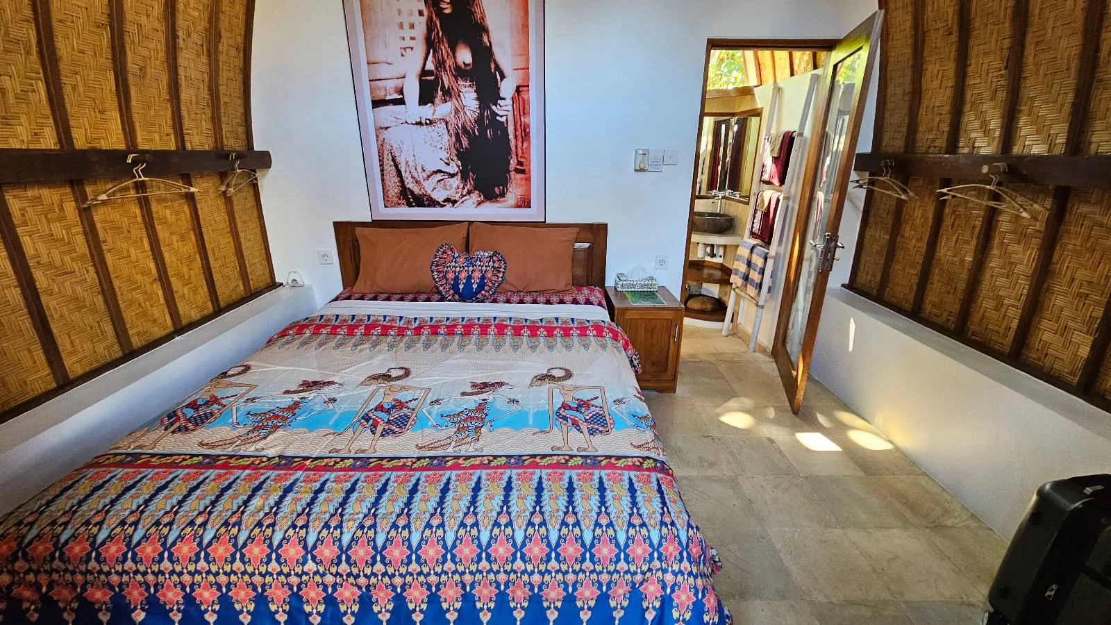 A cozy bedroom with a colorful, patterned bedspread on a double bed, wooden window shutters on both sides, a nightstand, wall art above the bed, and an open door leading to another room.