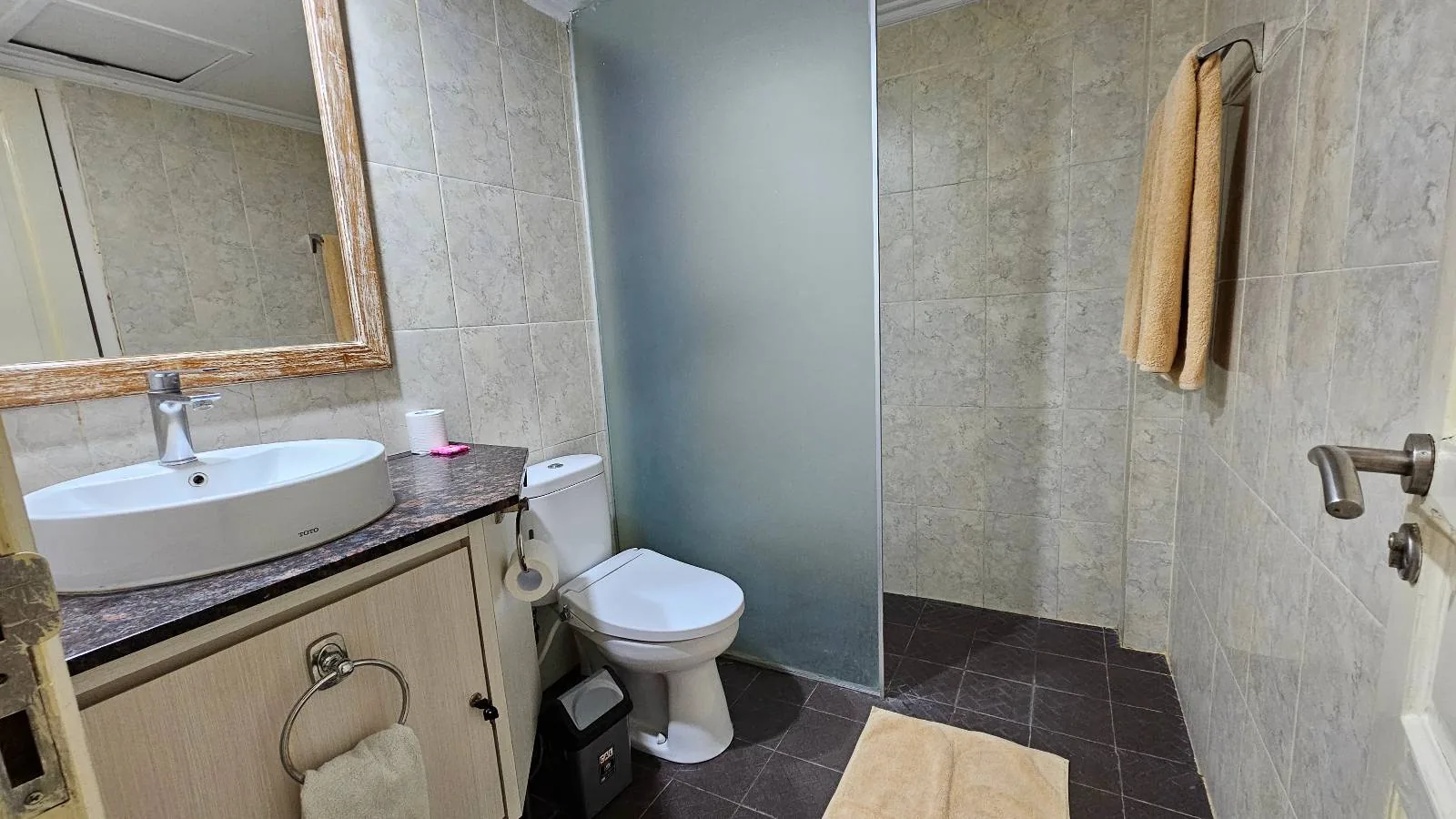 A modern bathroom with a sink and mirror on the left, a toilet in the center, and a glass shower partition on the right. A towel hangs on the wall and a mat is placed on the dark tiled floor.