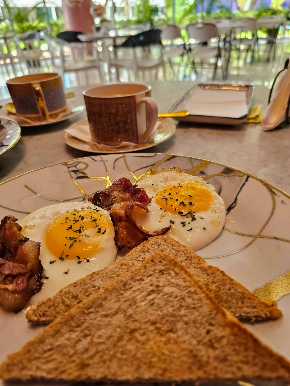 A breakfast setting with a plate of two sunny-side-up eggs sprinkled with herbs, pieces of bacon, and two slices of toasted bread. In the background, there is a cup of coffee and a sugar container on a reflective table.