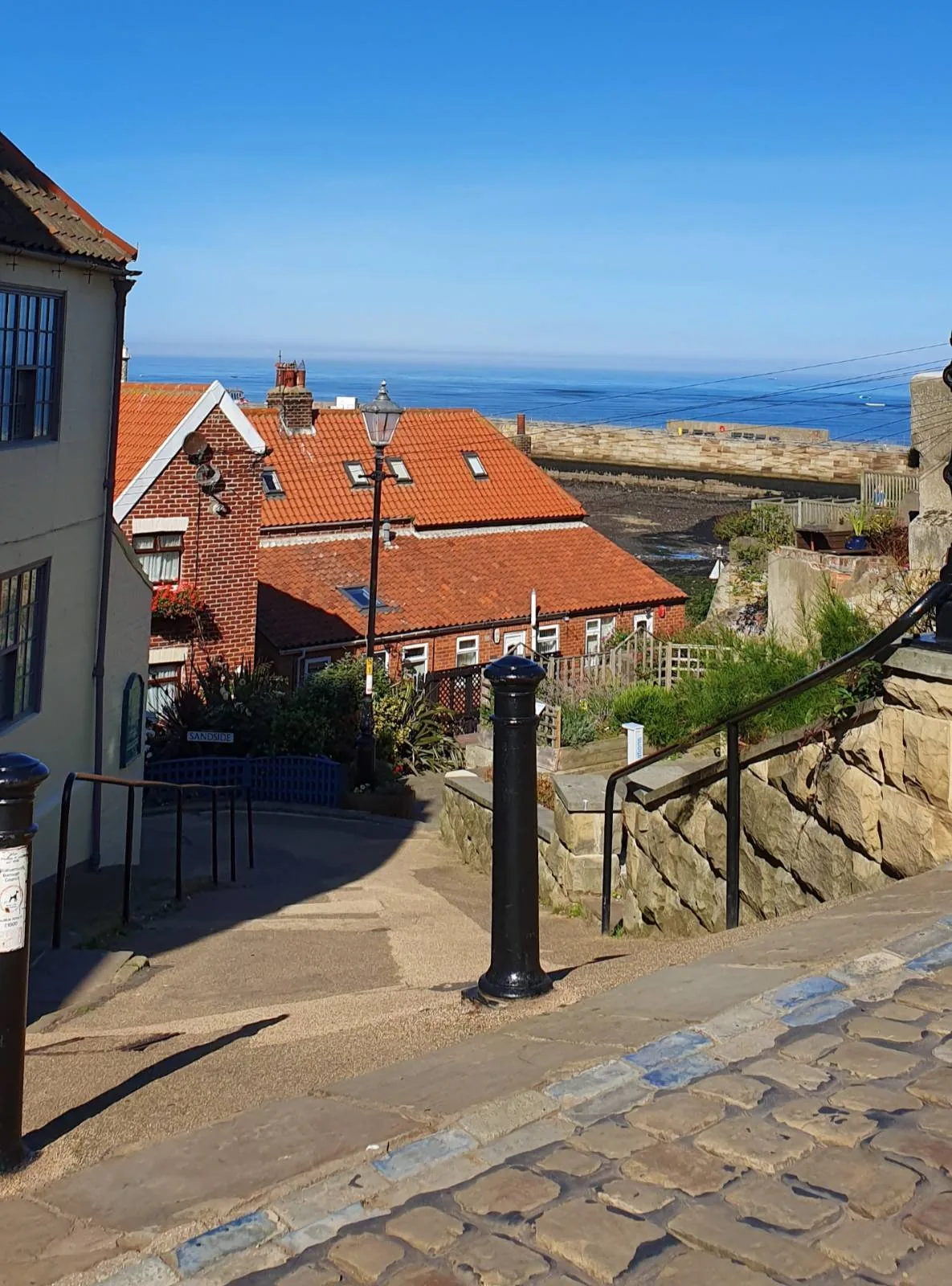 A coastal village scene with cobblestone paths leading down to houses with red roofs. A lamppost and stone walls line the path. The bright blue sea is visible in the background under a clear sky.
