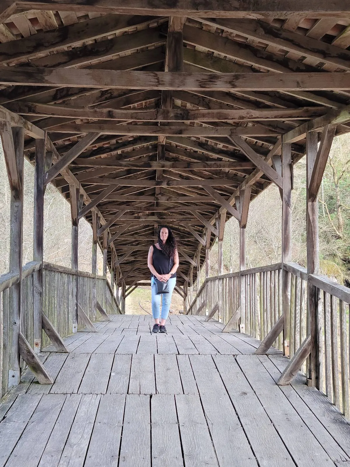 A person stands on a wooden covered bridge with a peaked roof, surrounded by trees. The bridge’s weathered planks and beams create a rustic atmosphere. The person is centered in the image, facing the camera.