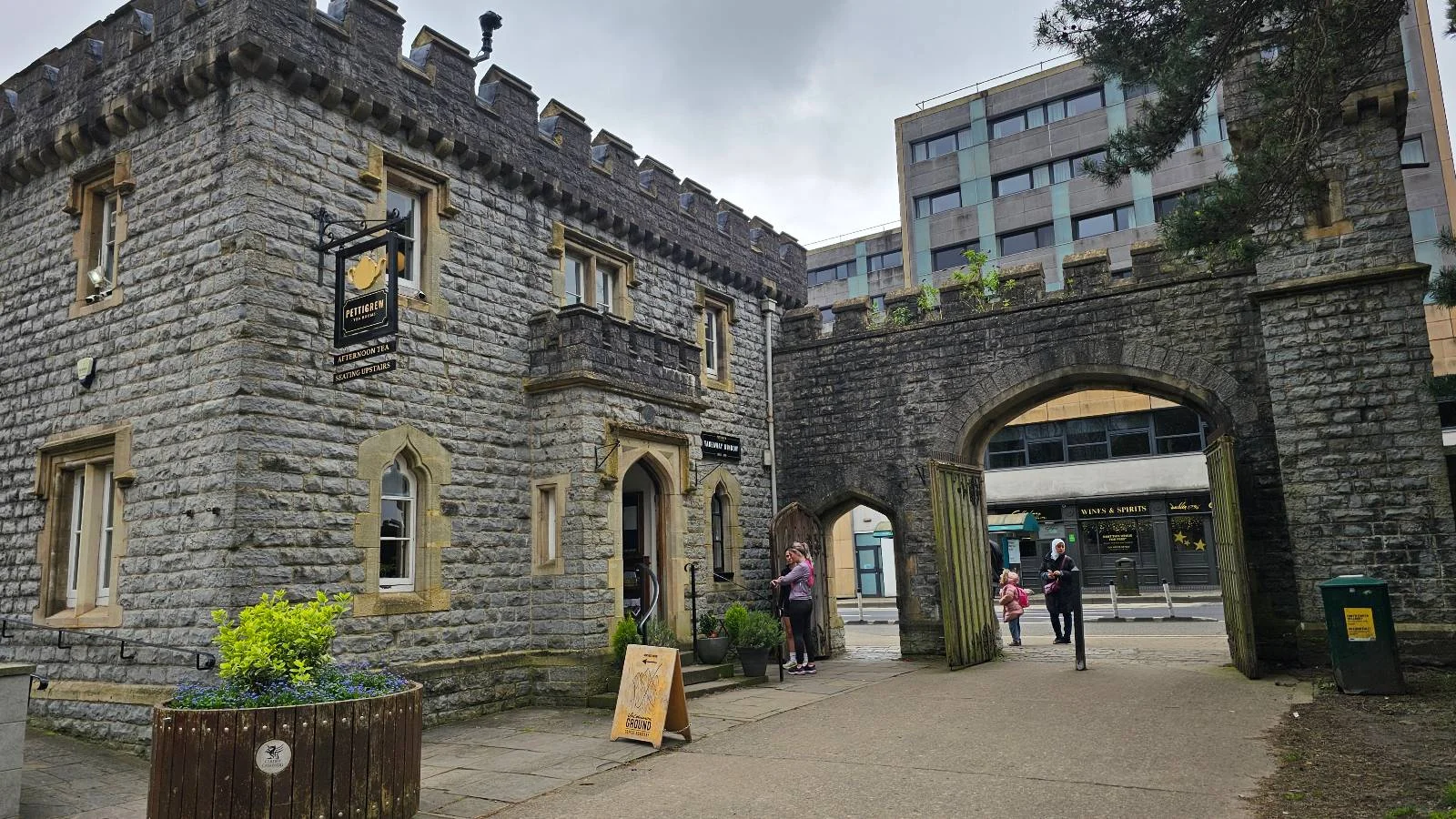 Old stone gatehouse with an arched entrance, juxtaposed with a modern building in the background, people walking by.