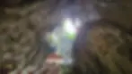 Inside a cave with a skylight above, stone stairs lead up to a building entrance. Green foliage is visible. A banner hangs over the entrance.