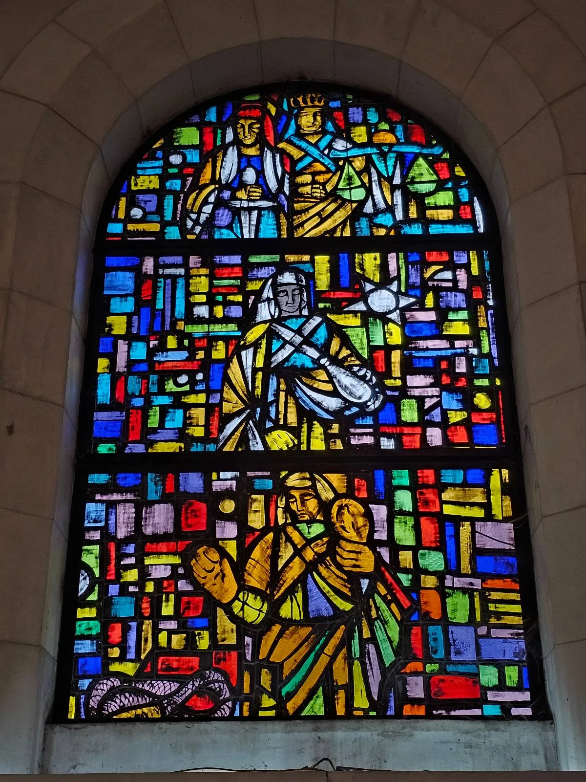 Stained glass window depicting religious figures in vibrant colors, including blue, yellow, red, and green. The design is detailed with geometric patterns surrounding the central figures in arches.