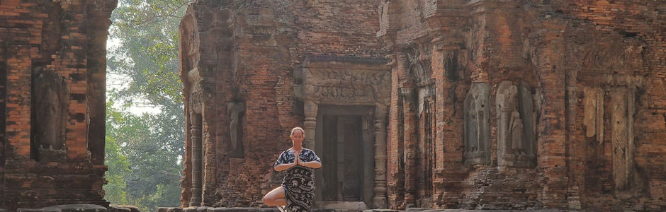 A person stands on one leg in a yoga pose in front of ancient, weathered brick temple ruins surrounded by trees and sunlight.