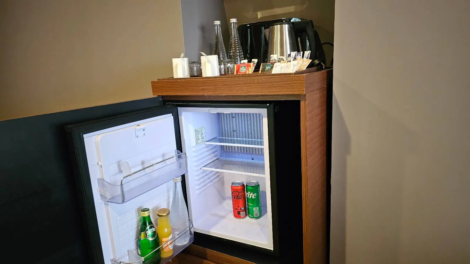 A small hotel minibar with an open fridge door reveals a few drinks, including a green bottle and cans. On top, there's a kettle and several cups, along with tea and coffee packets, neatly arranged.