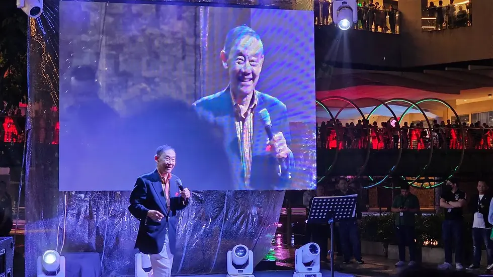Man in a suit, holding a microphone, stands on stage with large screen behind him. Crowd watching from balcony, colorful lights illuminate scene.
