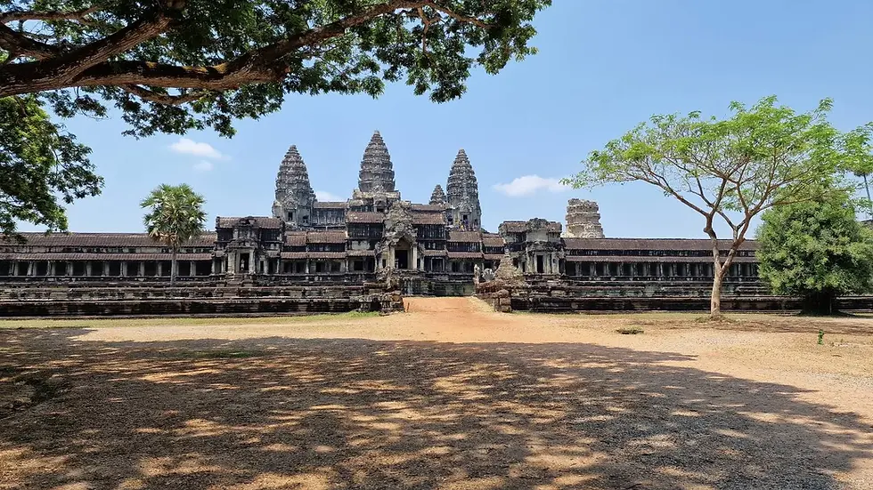 Angkor Wat temple with iconic spires under a clear blue sky, framed by green trees. Foreground shows shaded sandy ground.