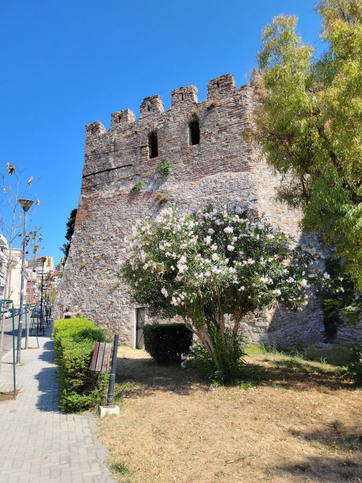 A stone fortress with crenellated walls stands under a clear blue sky, surrounded by green trees and bushes. A paved walkway and street lamps are visible on the left side.