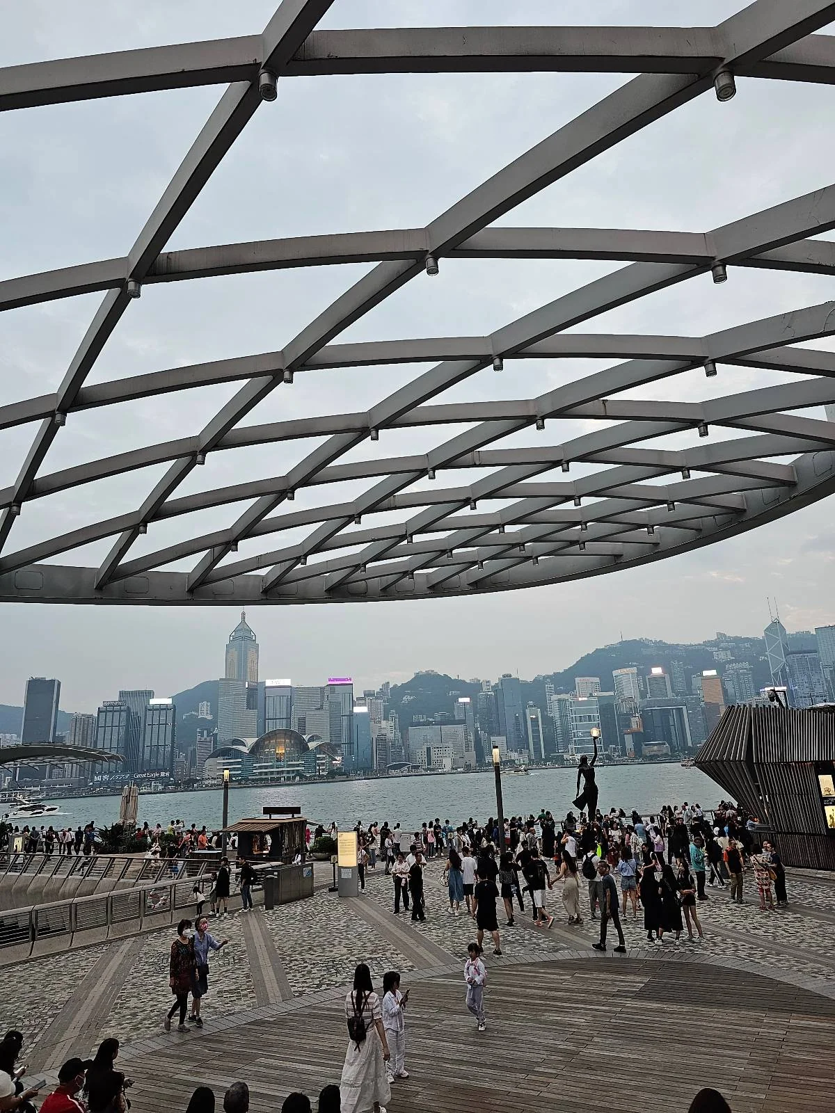 A crowd gathers at a waterfront under a modern, lattice-like canopy with a cityscape and mountains in the background.