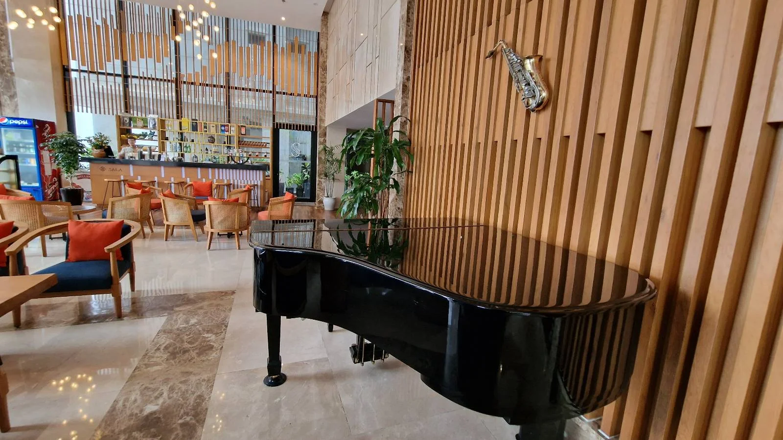 A sleek black grand piano is positioned in a modern, airy lobby. The space features wooden vertical slats, art on the wall, and contemporary furniture. A bar with stools and tables is visible in the background, with warm lighting throughout.