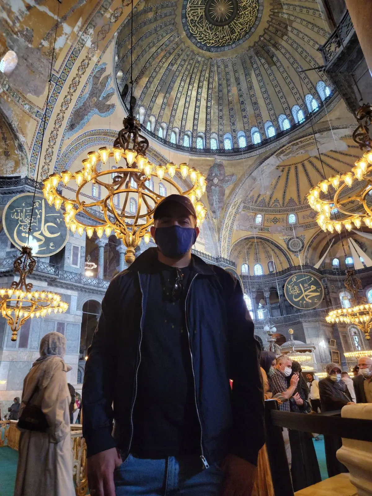 A person wearing a mask stands inside a large ornate building with chandeliers and a decorated domed ceiling.