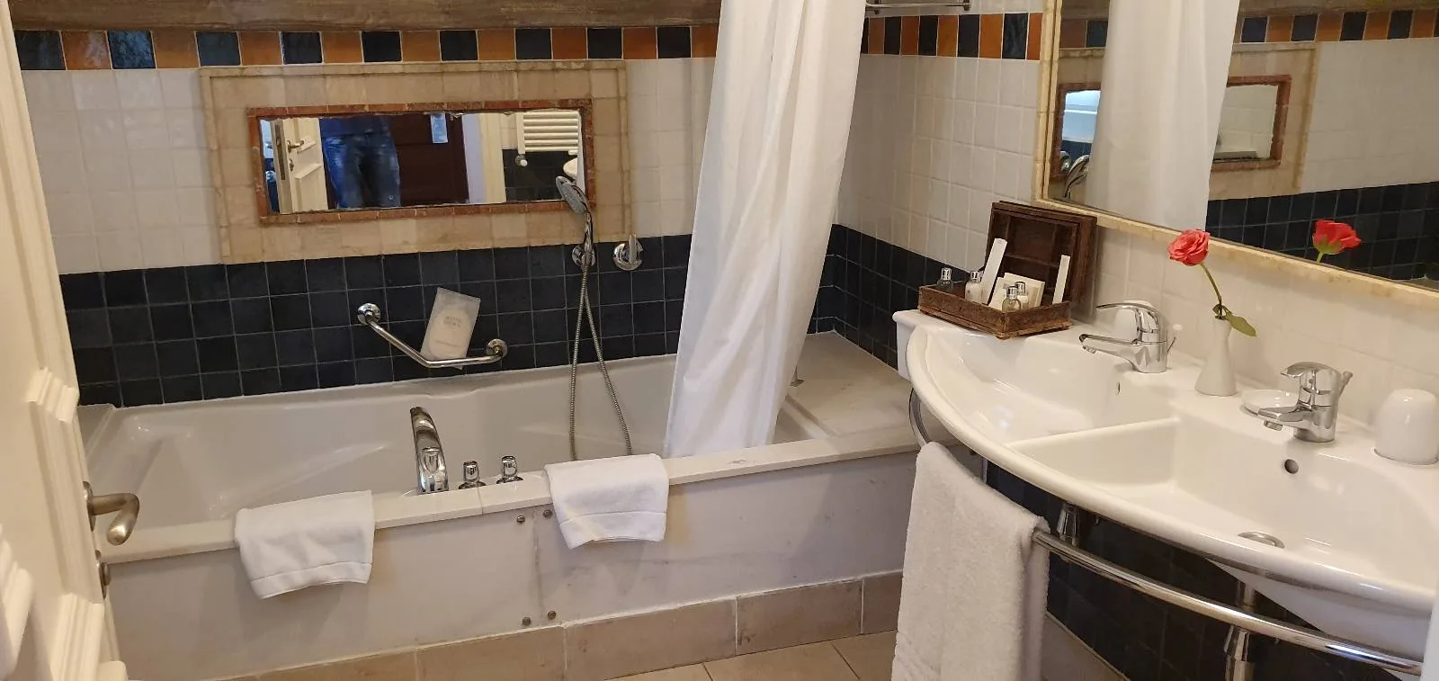 A bathroom with a bathtub featuring a wall-mounted showerhead and a curtain. Towels hang on the tub's edge. A large white sink is on the right, with toiletries and a vase with a red flower. A mirror is above the tub.