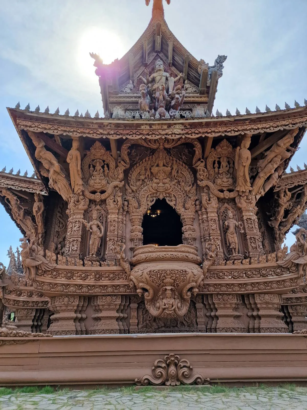 Intricately carved wooden temple with elaborate designs, viewed from the front, against a bright sky with the sun shining above the roof.