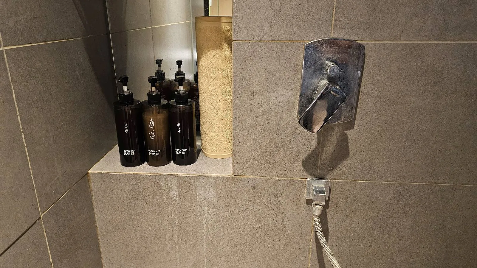 Four dark-colored bottles and one beige bottle are placed on a tiled shower shelf next to a metal shower control handle and attached hose against beige tiles.