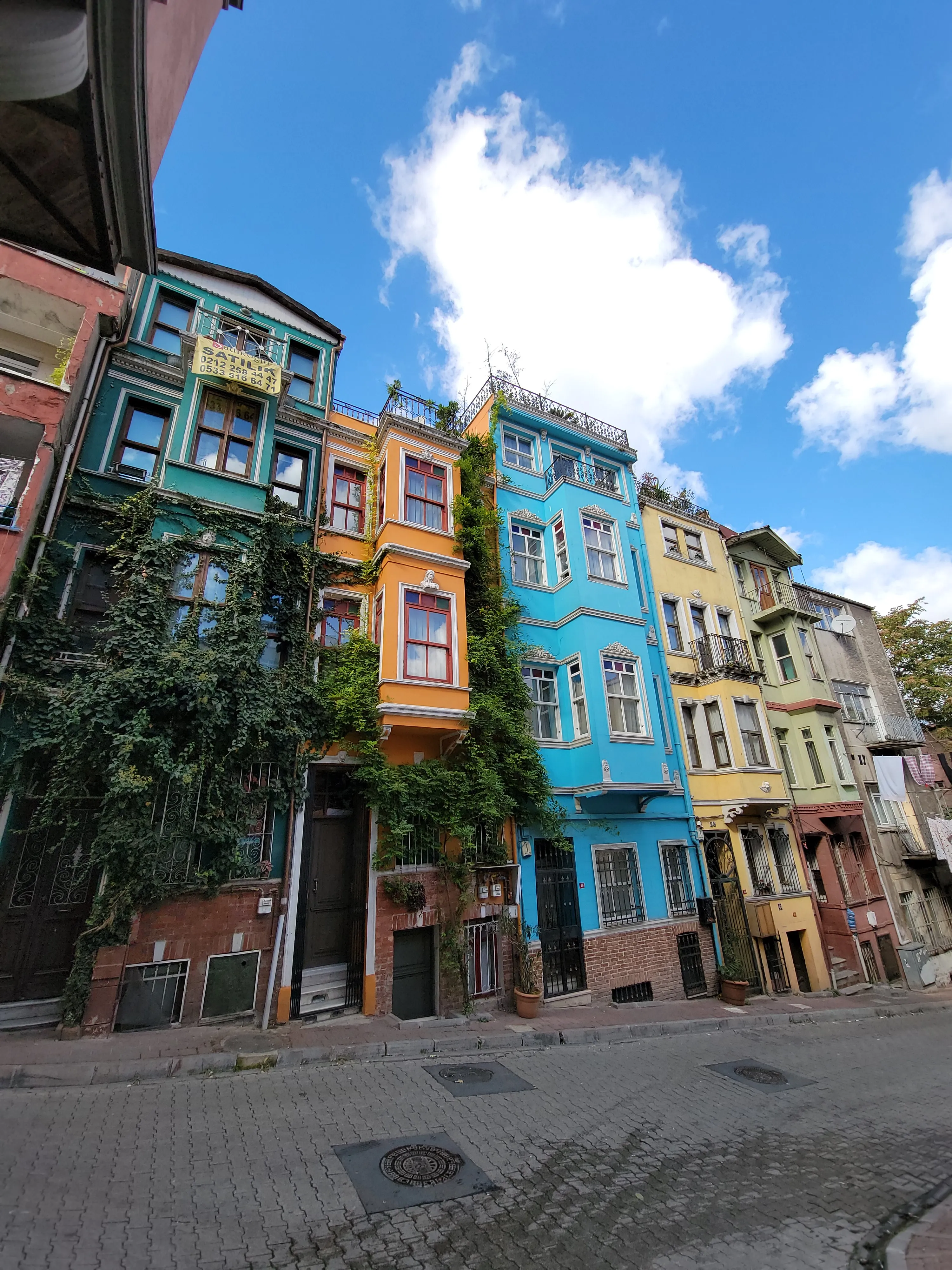 Colorful, narrow houses with ornate facades line a steep street under a bright blue sky with fluffy clouds. Green ivy climbs up one building, adding to the vibrant, lively urban scene.