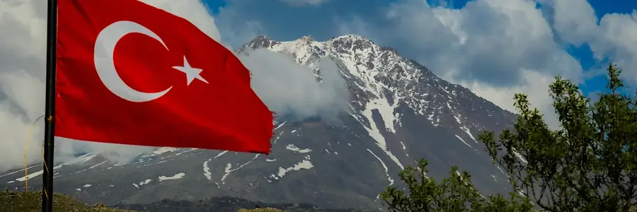 Panoramic view of rugged, snow-dusted mountains under a cloudy blue sky with the Turkish flag waving on the side, featured on the Türkiye travel guide.