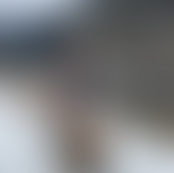 Two people pose on a wooden bench in a snowy landscape by a river, surrounded by pine trees and mountains in the background. One person stands while the other sits on the bench.