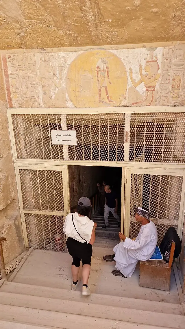 Two tourists approach the entrance of an ancient Egyptian tomb, where a seated person is stationed next to the gated doorway adorned with hieroglyphics and figures.